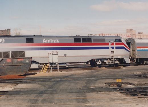 Side view of a silver Amtrak diesel locomotive numbered 809 with red and blue stripes, coupled to a silver baggage car, standing on a yard track behind paved crossings, a switch stand and a rusty dumpster.