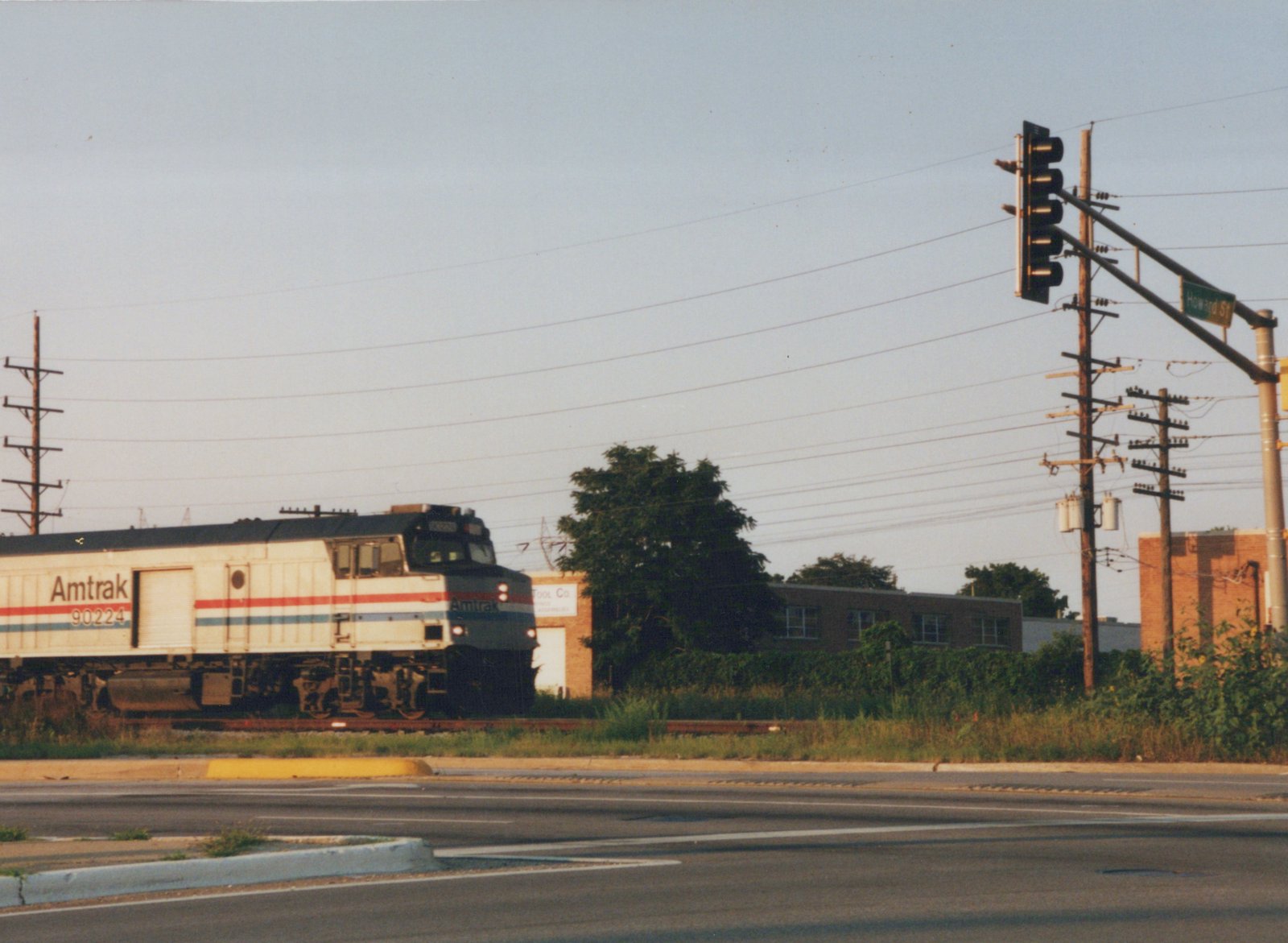 A silver Amtrak cab unit with red, white and blue stripes and a side roll-up door runs along tracks beside an empty street intersection with a traffic signal, utility poles and low brick industrial buildings.