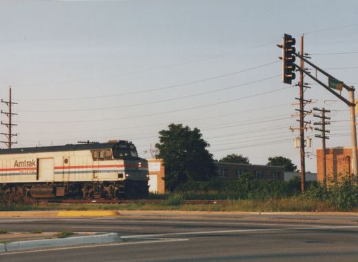 A silver Amtrak cab unit with red, white and blue stripes and a side roll-up door runs along tracks beside an empty street intersection with a traffic signal, utility poles and low brick industrial buildings.