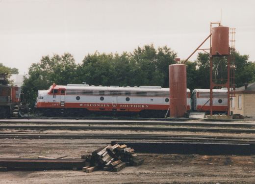 A silver and red streamlined passenger diesel lettered Wisconsin & Southern, number 10C, is coupled to a matching cabless unit. They stand in a yard behind several empty tracks, next to two red sand towers, with a pile of railroad ties in the foreground.