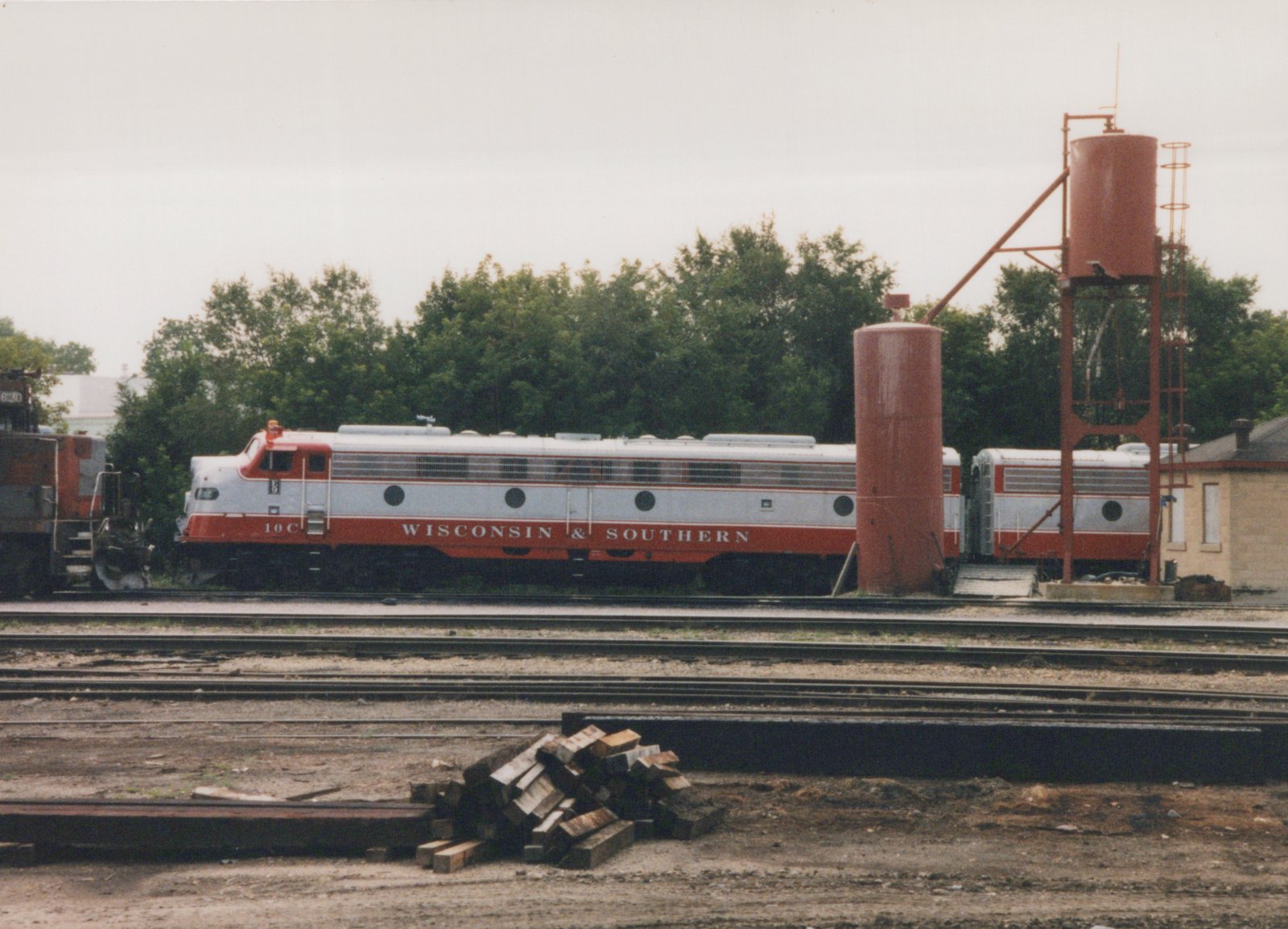 A silver and red streamlined passenger diesel lettered Wisconsin & Southern, number 10C, is coupled to a matching cabless unit. They stand in a yard behind several empty tracks, next to two red sand towers, with a pile of railroad ties in the foreground.
