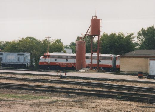 A red and silver passenger diesel locomotive set parked behind two red sand towers in a rail yard, with a blue-gray hood locomotive numbered 1792 at left and several empty tracks in the foreground.