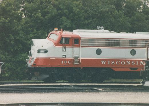 Side view of a red and light gray streamlined diesel locomotive numbered 10C, with WISCONSIN lettered on its side, standing on a track in front of dense green trees.
