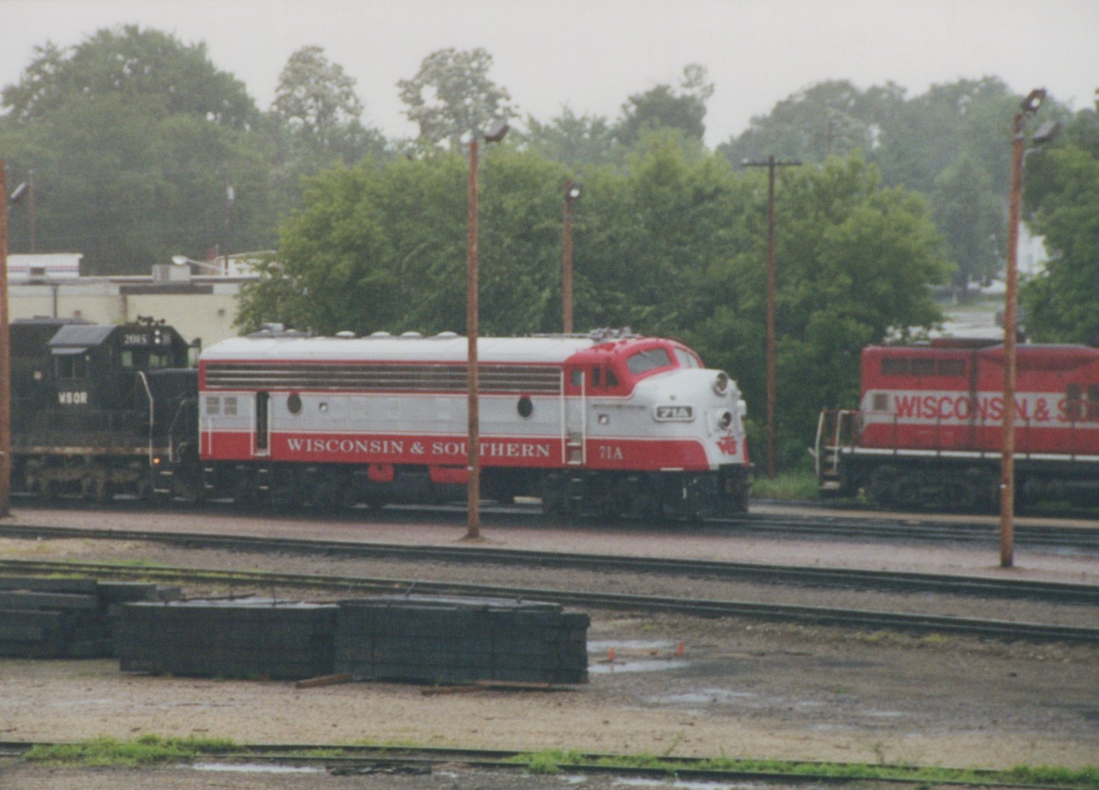 A red and silver streamlined diesel locomotive lettered Wisconsin & Southern, numbered 71A, stands in a rail yard on an overcast day, with a black locomotive behind it and a red locomotive at right.