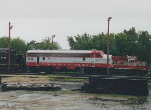 A red and silver streamlined diesel locomotive lettered Wisconsin & Southern and numbered 71A, shown from the side in a rain-soaked rail yard, with a black locomotive at left and a red and silver locomotive at right.