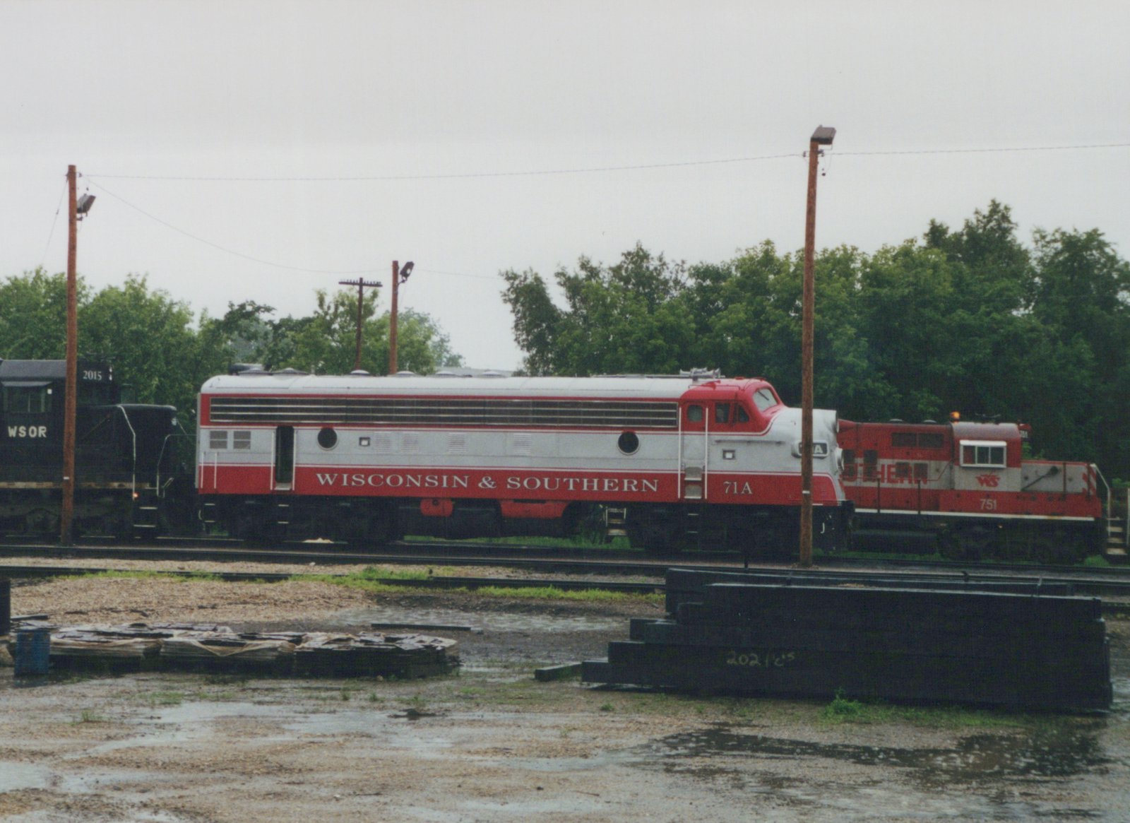 A red and silver streamlined diesel locomotive lettered Wisconsin & Southern and numbered 71A, shown from the side in a rain-soaked rail yard, with a black locomotive at left and a red and silver locomotive at right.