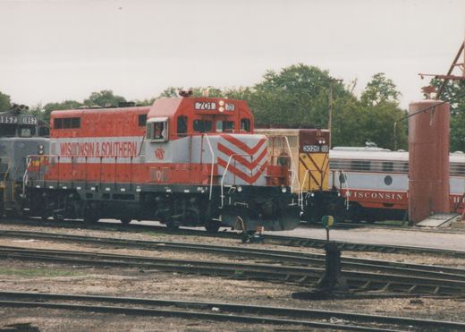 A red and gray Wisconsin and Southern diesel locomotive numbered 701 on a yard track, with a gray locomotive numbered 1852 behind it, a yellow and maroon locomotive numbered 3026, a gray and red passenger car lettered WISCONSIN, and a tall red tank.