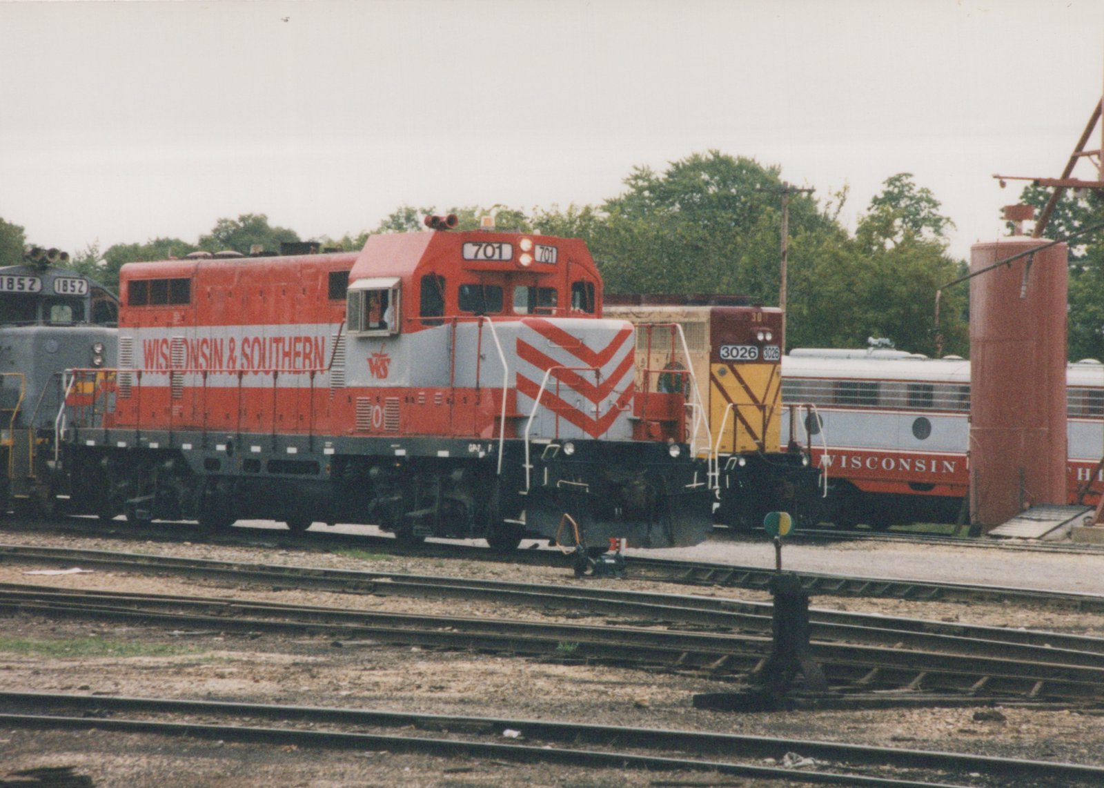 A red and gray Wisconsin and Southern diesel locomotive numbered 701 on a yard track, with a gray locomotive numbered 1852 behind it, a yellow and maroon locomotive numbered 3026, a gray and red passenger car lettered WISCONSIN, and a tall red tank.