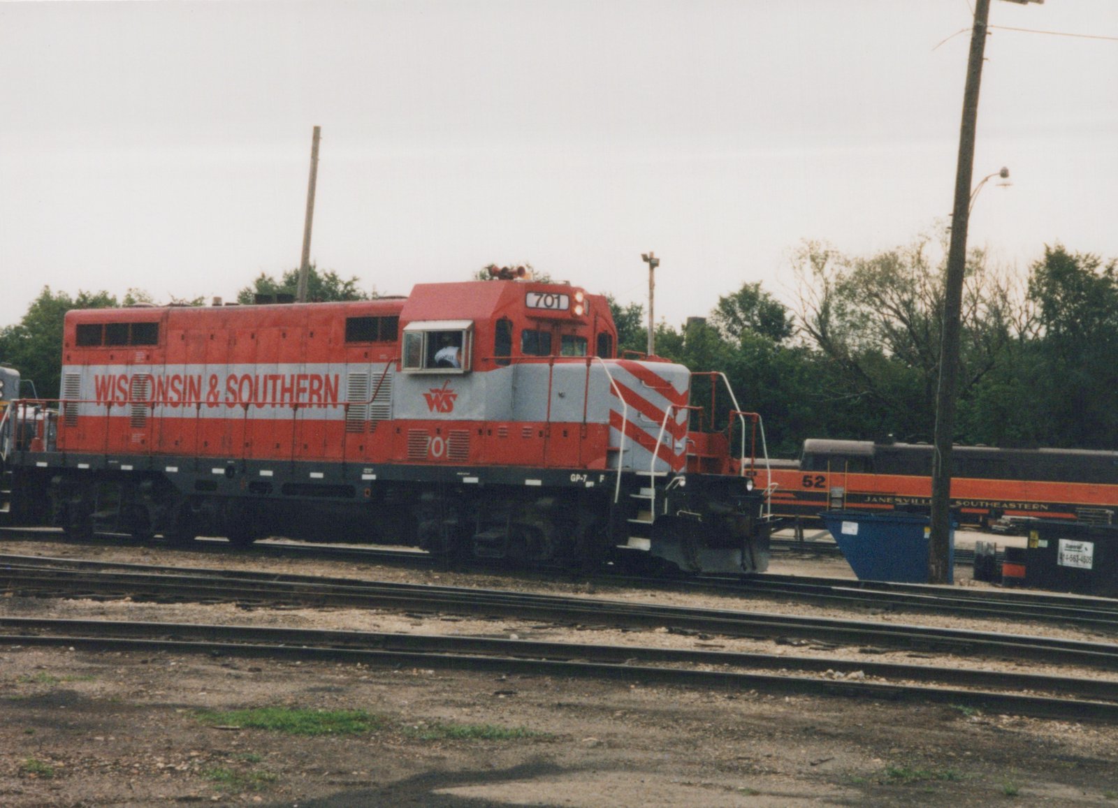 A red and gray diesel locomotive lettered Wisconsin & Southern and numbered 701 stands on a yard track under an overcast sky, with a dark car with an orange band behind it.