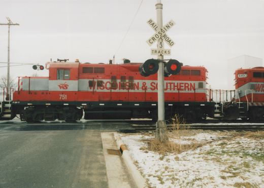 A red and silver Wisconsin & Southern diesel locomotive numbered 751 stands at a street crossing behind a crossbuck and flashing signals, with a second red locomotive numbered 4493 behind it and light snow on the ground.