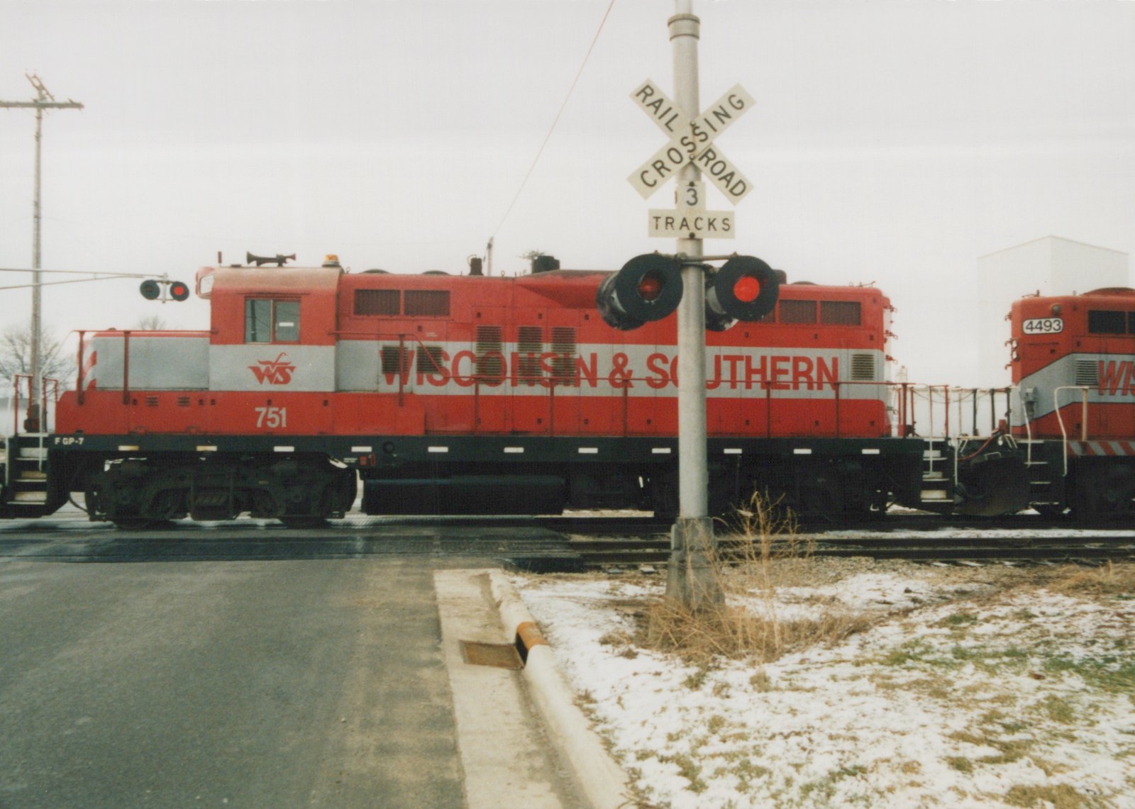 A red and silver Wisconsin & Southern diesel locomotive numbered 751 stands at a street crossing behind a crossbuck and flashing signals, with a second red locomotive numbered 4493 behind it and light snow on the ground.