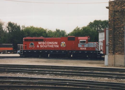 Side view of a red Wisconsin and Southern diesel locomotive numbered 1848, with a gray cab band and state seal emblem, standing beyond several yard tracks beside a brick building on an overcast day.