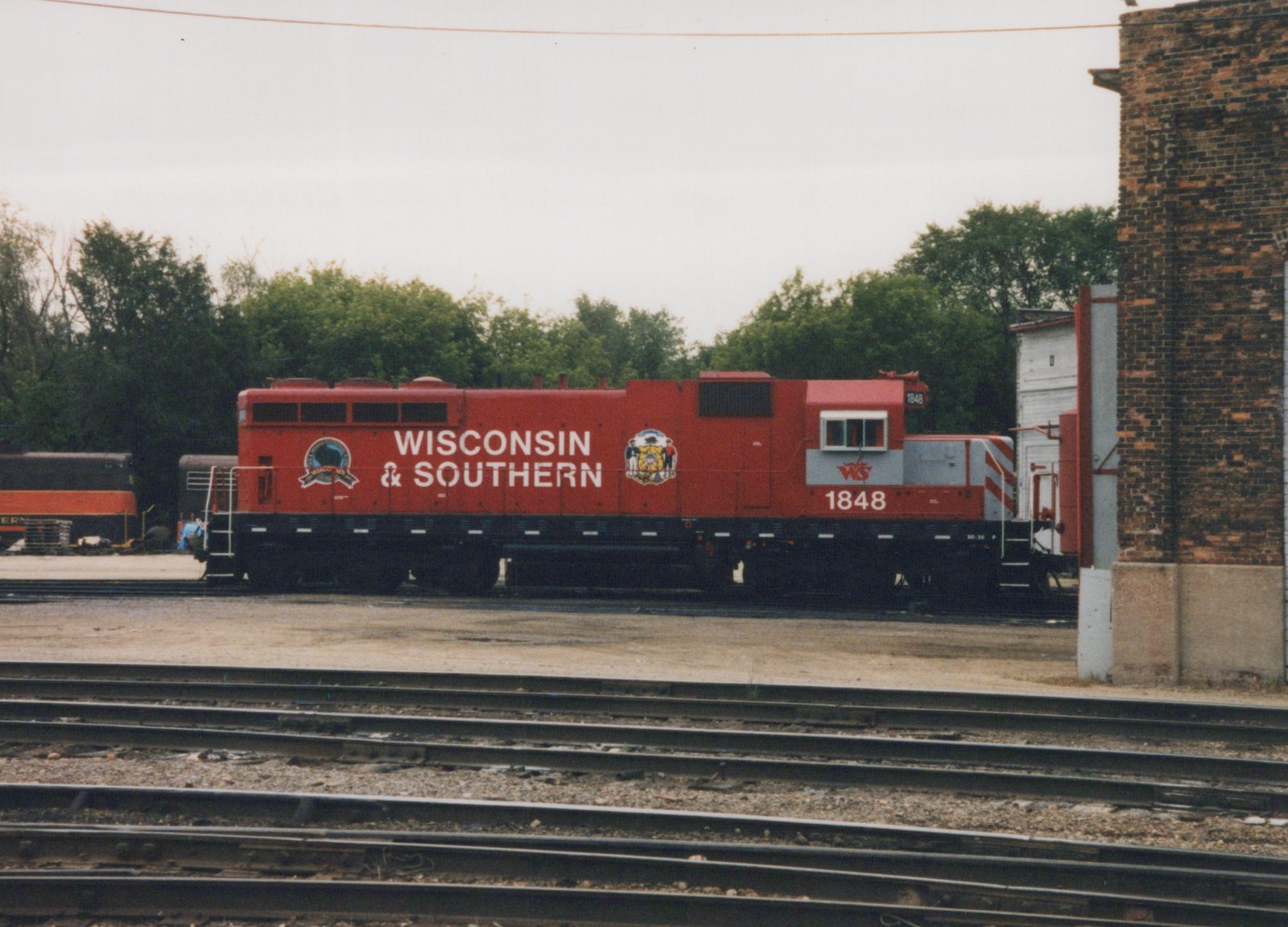 Side view of a red Wisconsin and Southern diesel locomotive numbered 1848, with a gray cab band and state seal emblem, standing beyond several yard tracks beside a brick building on an overcast day.