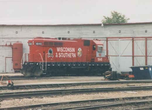 A red Wisconsin and Southern Railroad diesel locomotive numbered 1848 stands in front of a white enginehouse with numbered stalls, with empty yard tracks in the foreground.