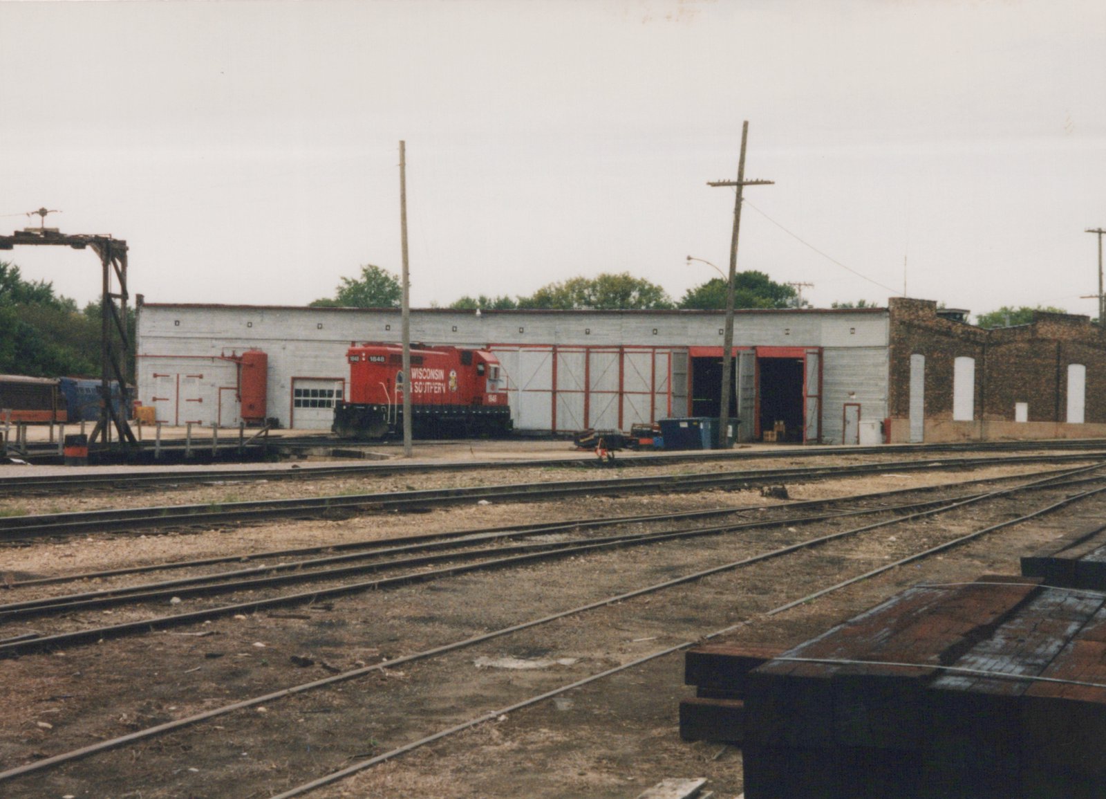 A red diesel locomotive stands in front of a long white roundhouse with red-framed doors, with yard tracks and a stack of ties in the foreground under an overcast sky.