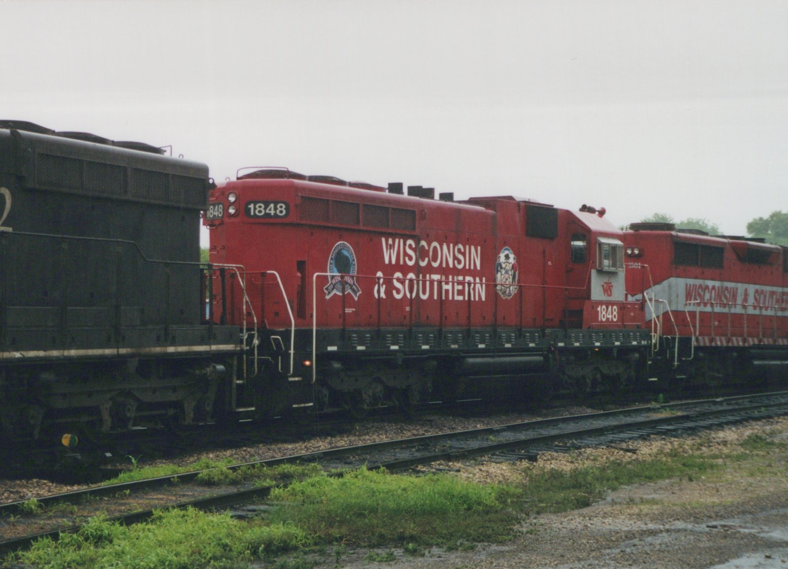 A red diesel locomotive lettered Wisconsin & Southern and numbered 1848 stands on a track, coupled to a black locomotive on the left and a red-and-grey locomotive on the right, with grass and a wet gravel area in front.