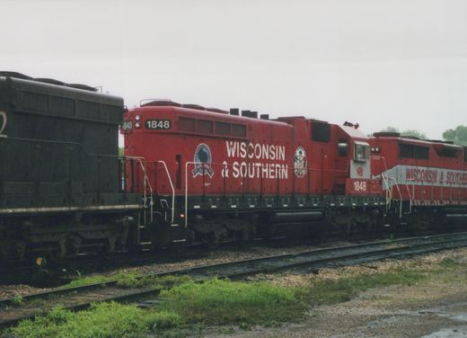 A red diesel locomotive lettered Wisconsin & Southern and numbered 1848 stands on a track, coupled to a black locomotive on the left and a red-and-grey locomotive on the right, with grass and a wet gravel area in front.