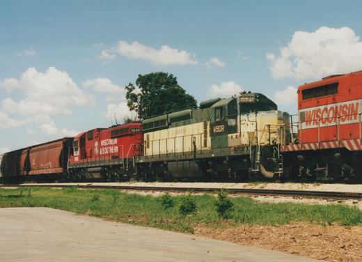 A red locomotive lettered Wisconsin & Southern and a weathered green and cream locomotive numbered 6625 stand on a track with brown covered hoppers behind, next to an orange locomotive at right, under a blue sky with clouds.