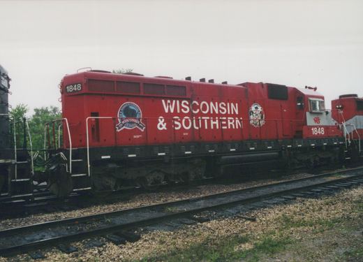 Side view of a red diesel locomotive numbered 1848, lettered Wisconsin & Southern with a round sesquicentennial emblem and the Wisconsin state seal, coupled between a black locomotive and another red locomotive on a track under an overcast sky.