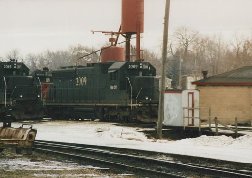 A dark diesel locomotive numbered 2009 and lettered WSOR sits beneath a red sand tower in a snowy rail yard, with two more locomotives at left and a small white shed at right.