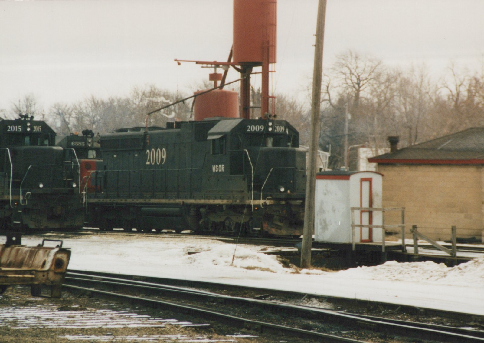 A dark diesel locomotive numbered 2009 and lettered WSOR sits beneath a red sand tower in a snowy rail yard, with two more locomotives at left and a small white shed at right.