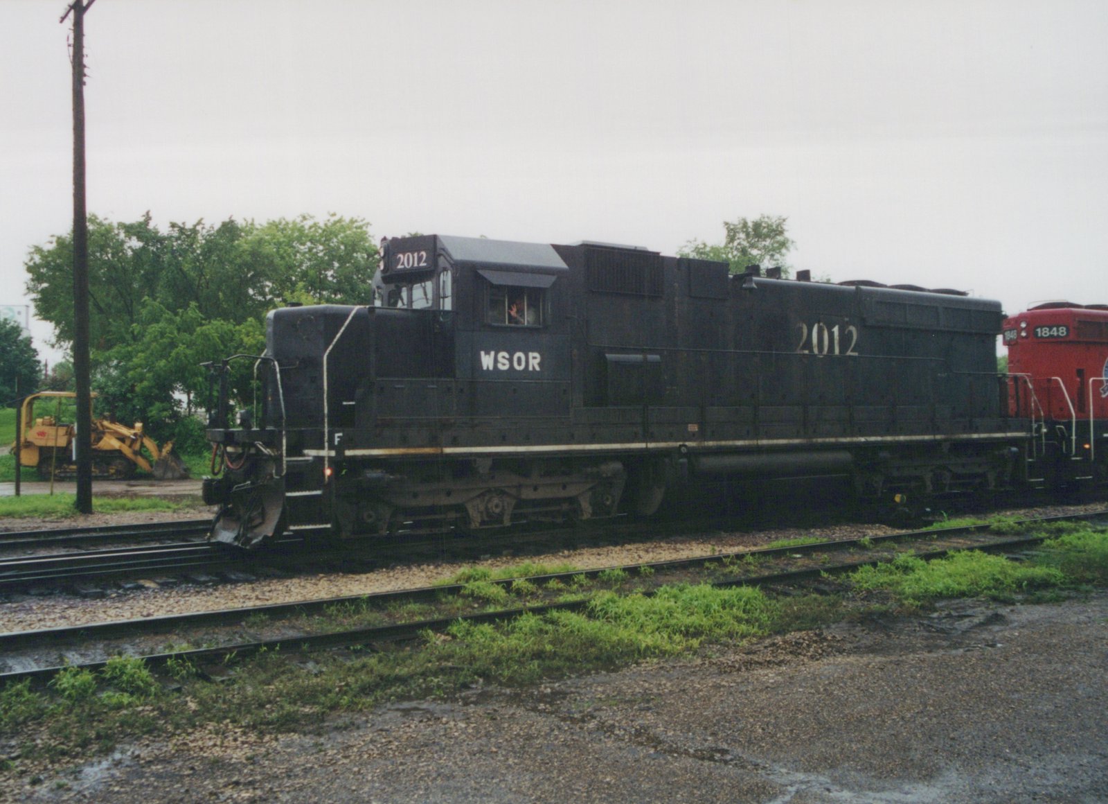 A black diesel locomotive lettered WSOR and numbered 2012 rolls along the tracks with a red locomotive numbered 1848 behind it. A crew member waves from the cab window, and trees and a yellow construction machine stand in the background.