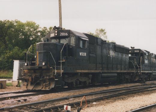 Two black four-axle diesel locomotives coupled together on a yard track under a gray sky, the lead unit numbered 2016 and lettered WSOR, with trees behind them.