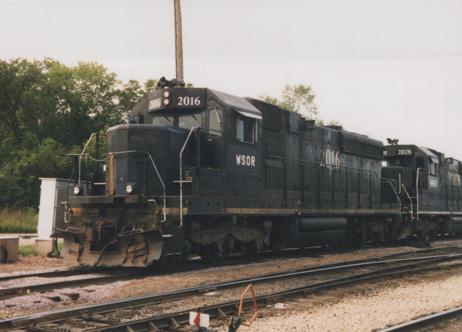 Two black four-axle diesel locomotives coupled together on a yard track under a gray sky, the lead unit numbered 2016 and lettered WSOR, with trees behind them.