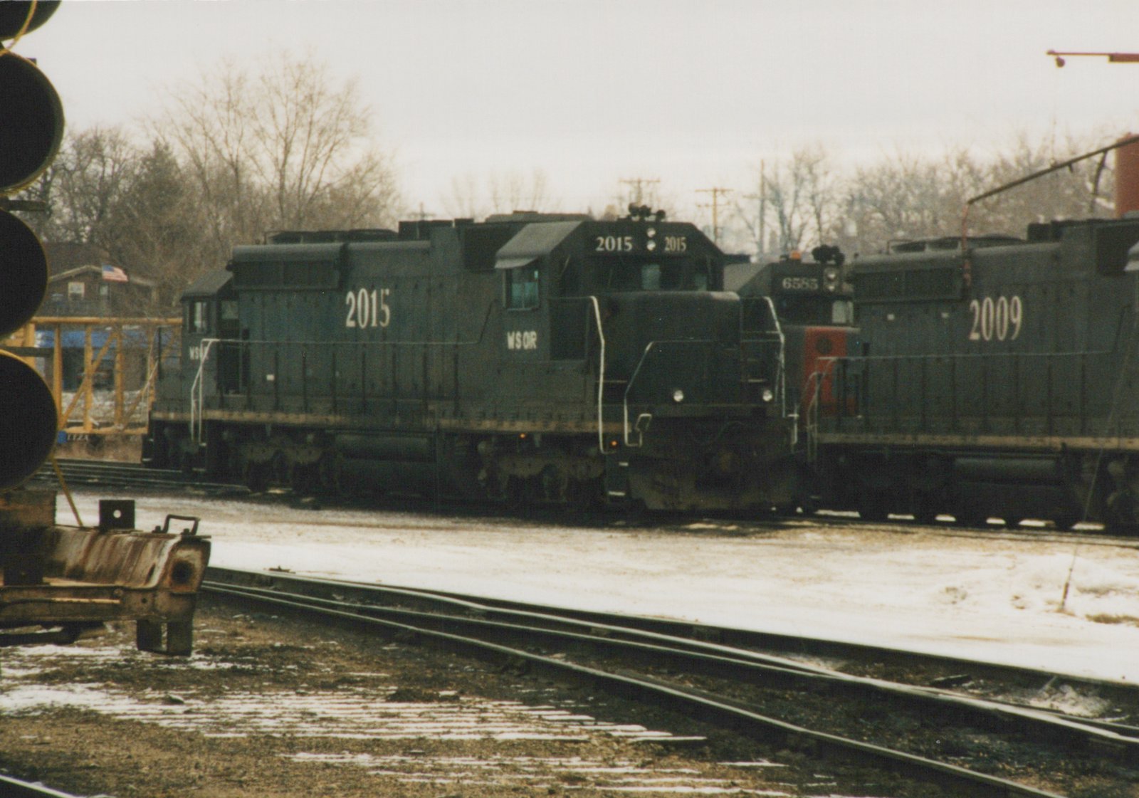A black diesel locomotive numbered 2015 and lettered WSOR stands on a yard track in light snow, with a locomotive with a red nose and another numbered 2009 behind it, bare trees and an overcast sky beyond.