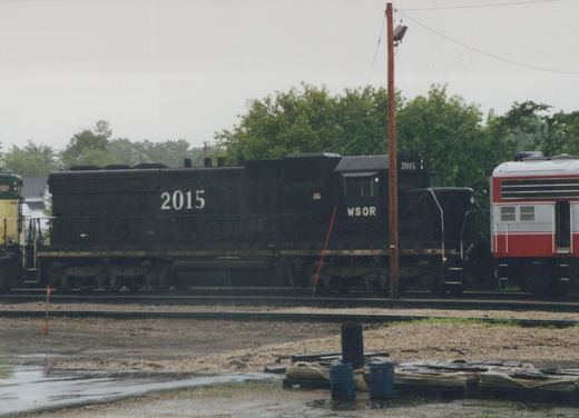 A black six-axle diesel locomotive lettered WSOR and numbered 2015, seen from the side on a yard track, with part of a yellow and green locomotive at the left, a silver and red passenger car at the right, and wet ground and stacked materials in front.