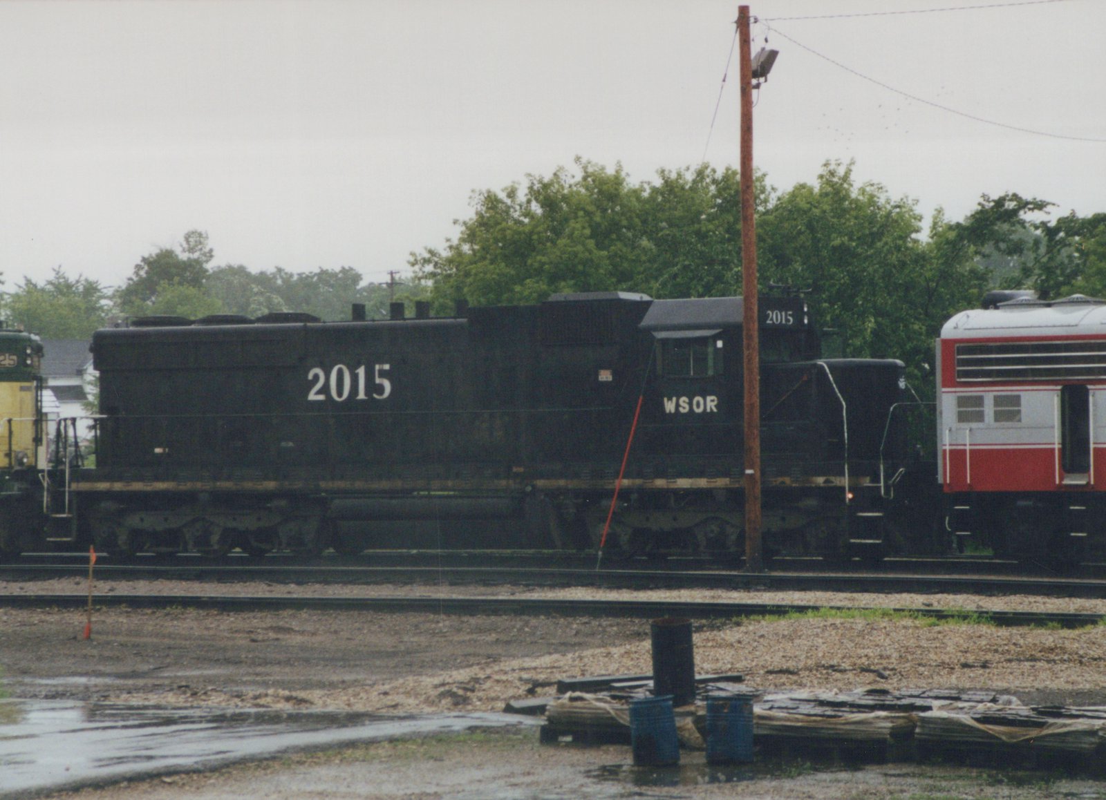 A black six-axle diesel locomotive lettered WSOR and numbered 2015, seen from the side on a yard track, with part of a yellow and green locomotive at the left, a silver and red passenger car at the right, and wet ground and stacked materials in front.