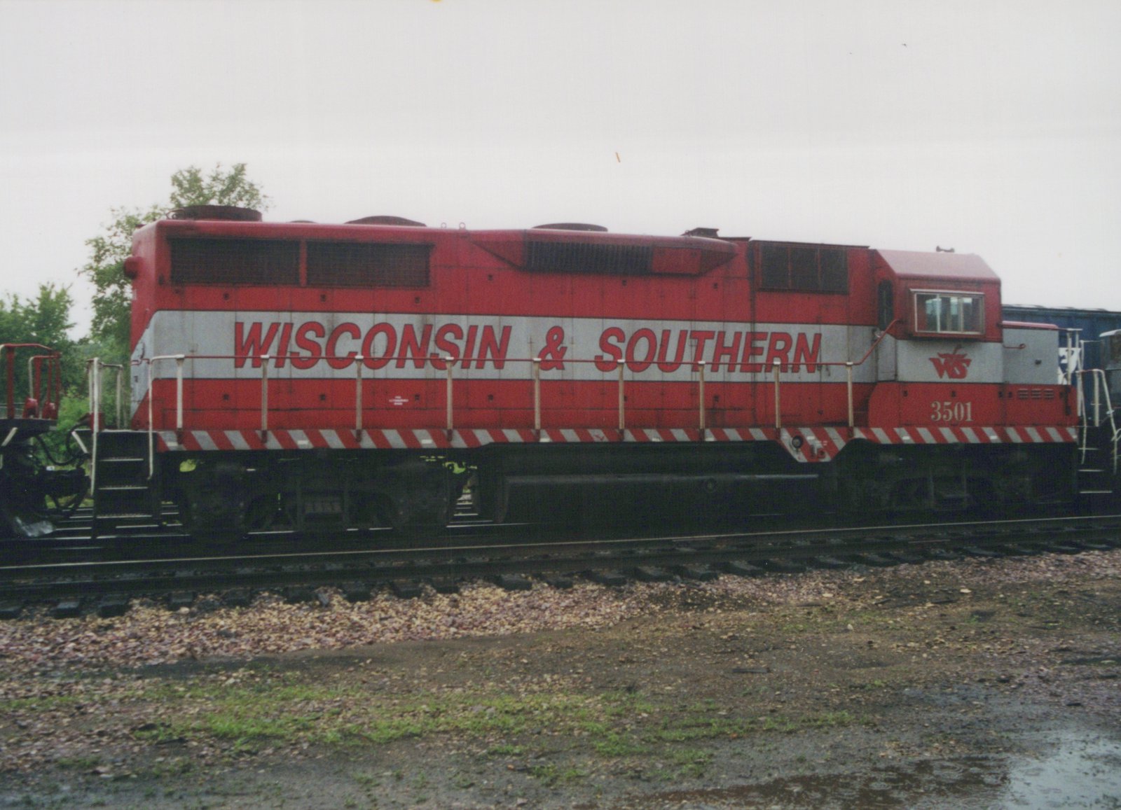 Side view of a red and silver Wisconsin & Southern diesel locomotive numbered 3501 on a track, with wet gravel and puddles in the foreground and trees behind it.