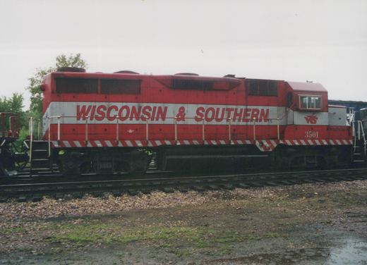 Side view of a red and silver Wisconsin & Southern diesel locomotive numbered 3501 on a track, with wet gravel and puddles in the foreground and trees behind it.