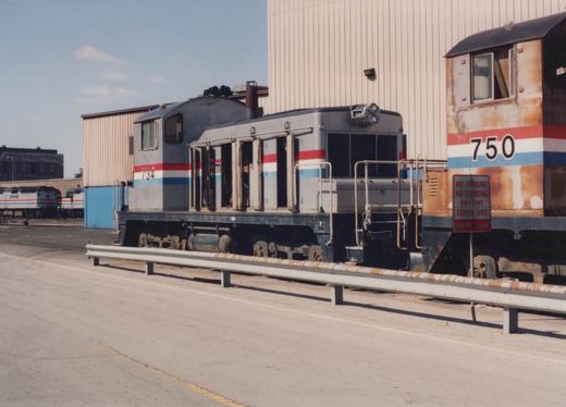 A gray switcher locomotive with red, white and blue stripes and its engine doors open stands beside a rusty switcher numbered 750, behind a guardrail in front of a tall metal building.