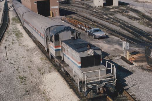 Seen from above, a silver Amtrak switcher with red, white and blue stripes is coupled to three stainless steel passenger cars beside a brick shop building, a row of wheelsets and a parked blue car.