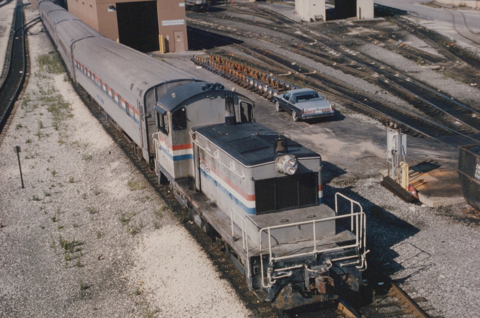 Seen from above, a silver Amtrak switcher with red, white and blue stripes is coupled to three stainless steel passenger cars beside a brick shop building, a row of wheelsets and a parked blue car.