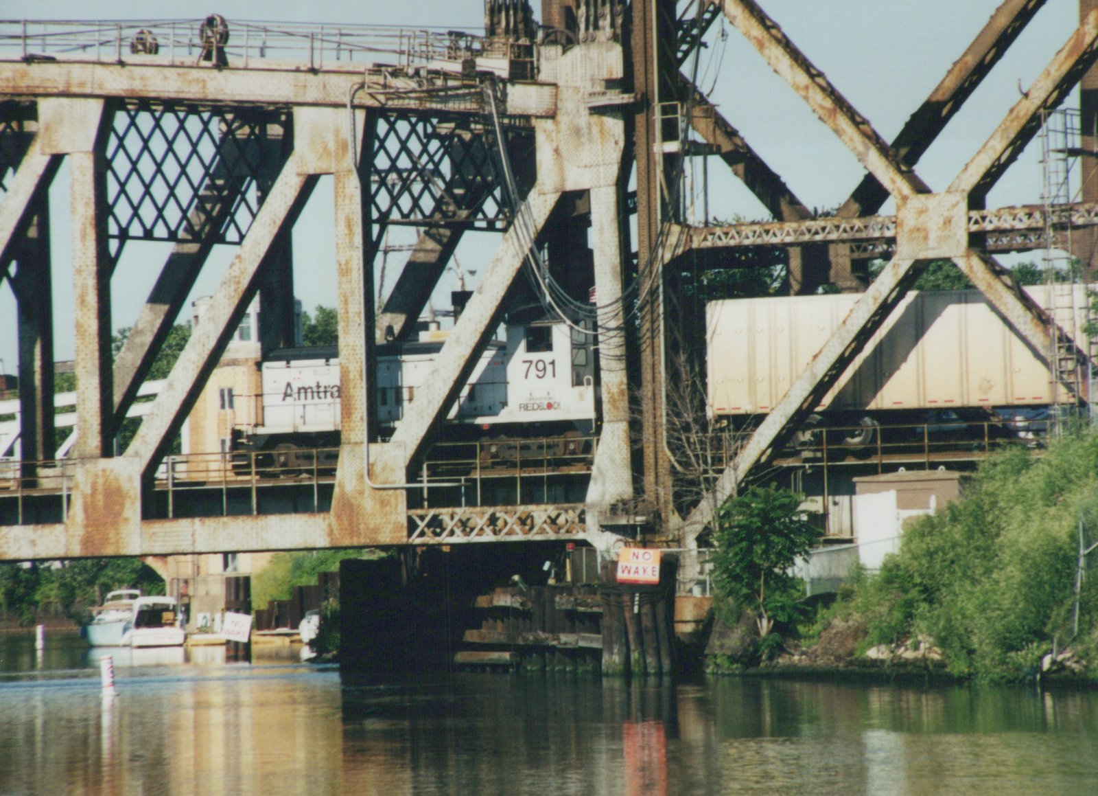 A gray and white Amtrak locomotive numbered 791 and a white trailer car seen through the riveted truss of a railroad bridge above the river, with NO WAKE signs on the pier and on the shore.