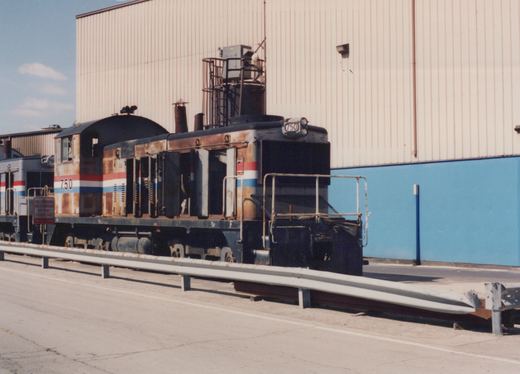 A rusted gray end-cab diesel switcher numbered 750, with red, white and blue stripes and its hood doors open, stands behind a roadside guardrail in front of a tall beige and blue building, with a silver locomotive behind it.