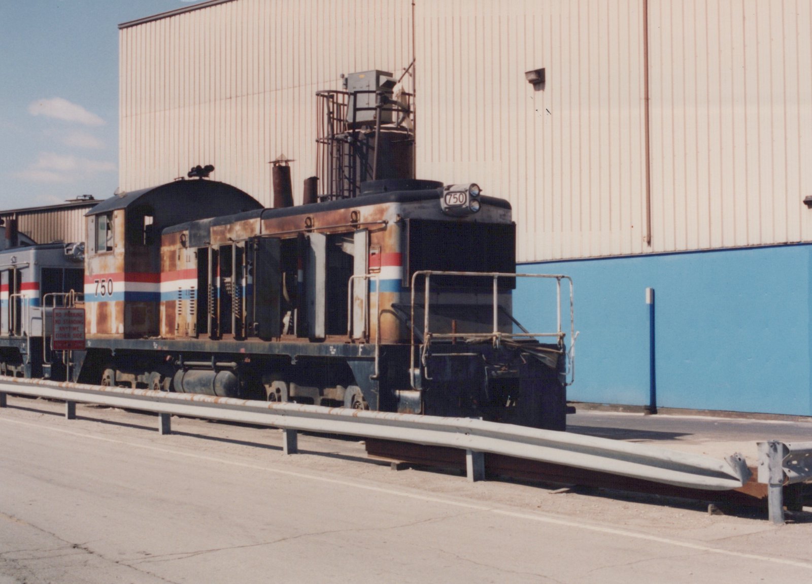 A rusted gray end-cab diesel switcher numbered 750, with red, white and blue stripes and its hood doors open, stands behind a roadside guardrail in front of a tall beige and blue building, with a silver locomotive behind it.