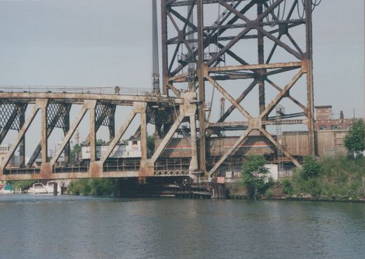 A rusty steel truss railroad bridge with a tall lift tower over a river, with a white Amtrak switcher numbered 794 and striped cars crossing behind the truss.
