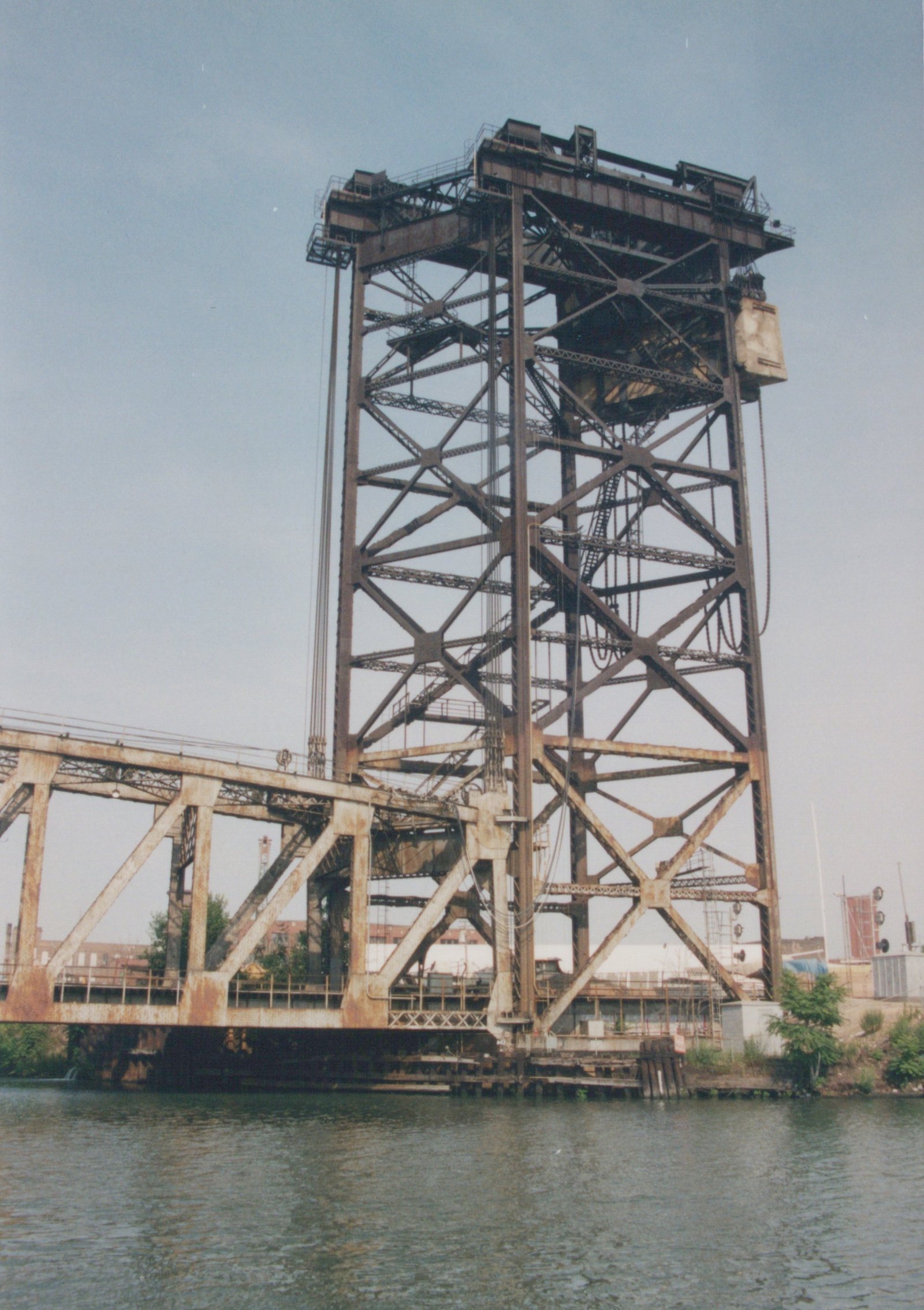 A rusty, steel vertical-lift bridge over a river, with its lift tower and counterweight visible against a pale sky.