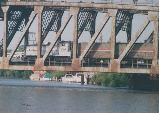 A white Amtrak switcher numbered 794 and a striped Amtrak car with graffiti seen through the riveted diagonal members of a rusted truss bridge, with moored boats and ducks on the river below.