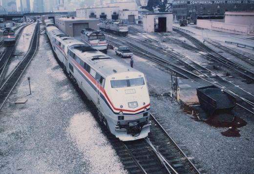 An elevated view of a silver Amtrak diesel locomotive with red, white and blue stripes, numbered 823, leading a train through a rail yard. Several more Amtrak locomotives, service buildings, a pickup truck and a brick warehouse signed Continental Paper Grading Co. are behind it, and a city skyline r
