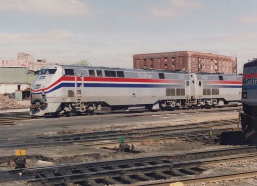 Two silver Amtrak diesel locomotives with red, white and blue stripes stand coupled on a yard track, with switches in the foreground and an old brick warehouse behind them.