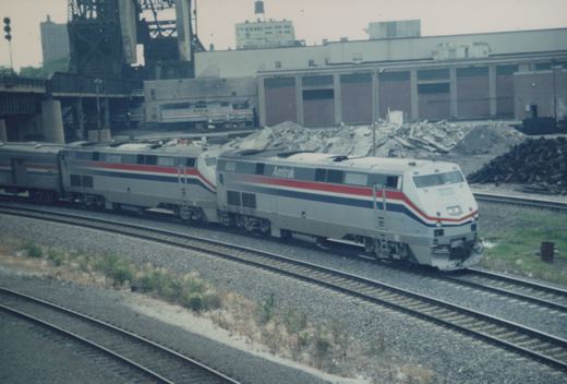 Two silver Amtrak diesel locomotives with red, white and blue stripes pull a train around a curve past rubble piles, a brick building and a tall steel lift bridge.