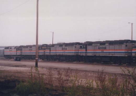Four silver locomotives with red, white and blue stripes, lettered Amtrak, parked in a row behind a gravel road, a utility pole and weeds under a grey sky.