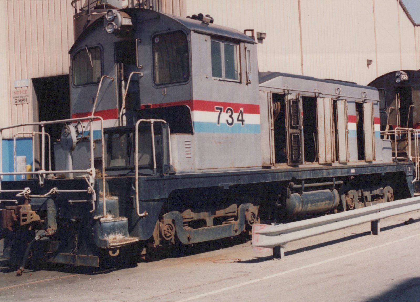A small gray end-cab diesel switcher numbered 734, with a red, white and blue stripe and its hood doors open, parked beside a shop building with another locomotive, number 750, behind it.