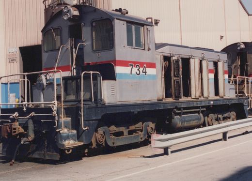 A small gray end-cab diesel switcher numbered 734, with a red, white and blue stripe and its hood doors open, parked beside a shop building with another locomotive, number 750, behind it.