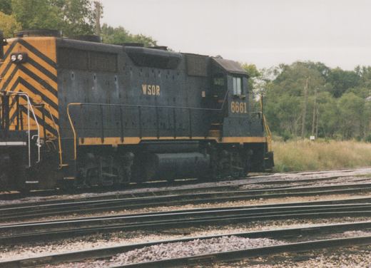 Side view of a dark blue-black diesel locomotive lettered WSOR and numbered 6661, with yellow trim and yellow-and-black chevrons on its rear end, standing on one of several tracks beside a grassy field and trees.