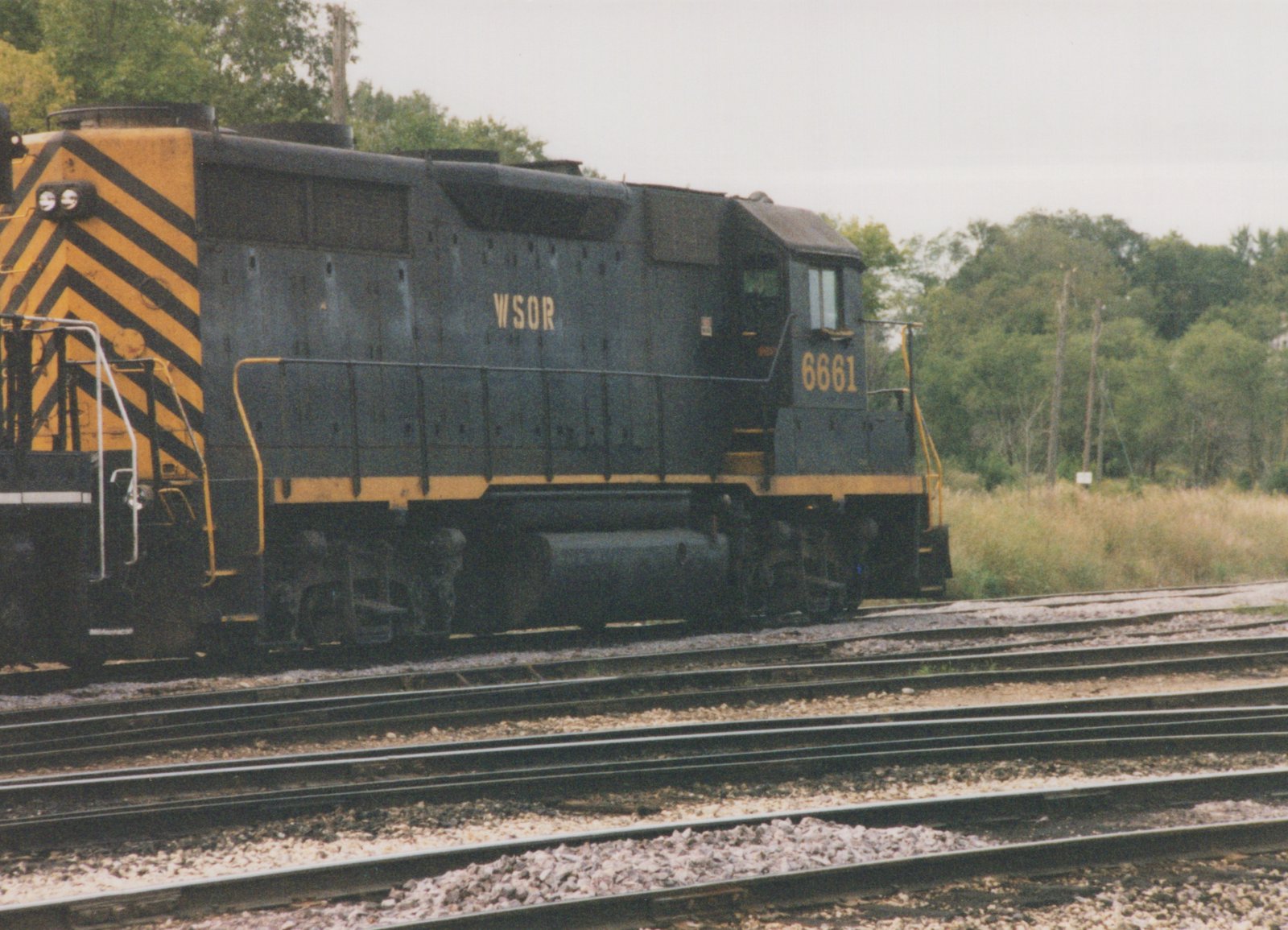 Side view of a dark blue-black diesel locomotive lettered WSOR and numbered 6661, with yellow trim and yellow-and-black chevrons on its rear end, standing on one of several tracks beside a grassy field and trees.